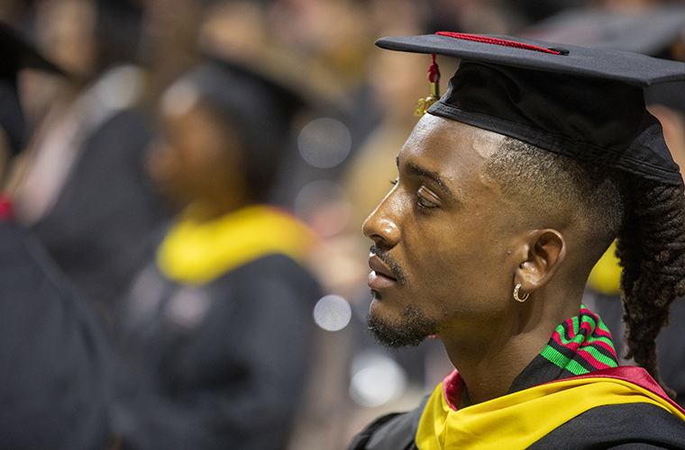Person looks forward in black graduation gown and cap with yellow and red collar. Other graduates are seated in the background